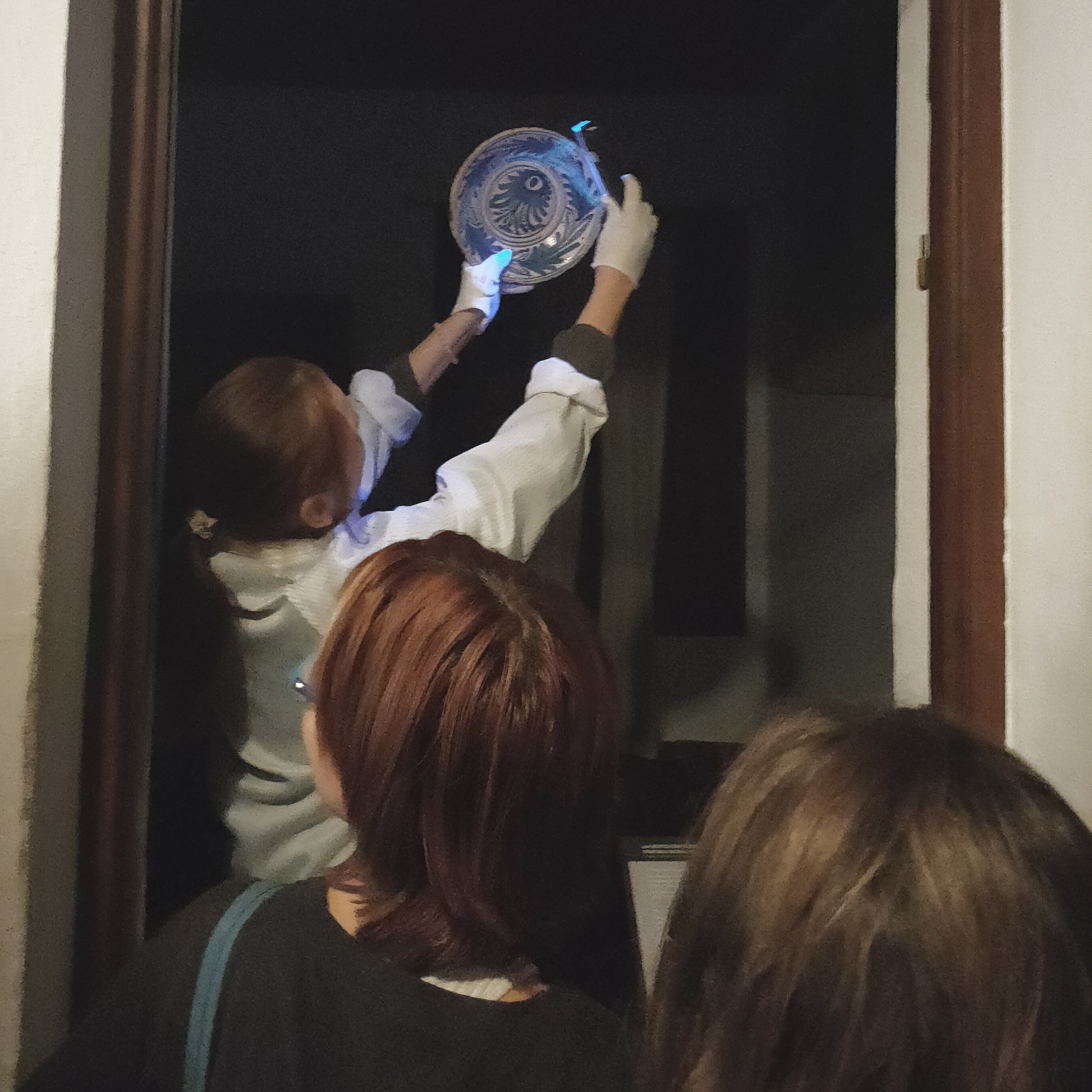 En una estancia interior, una especialista muestra a varias personas cómo la luz azul puede revelar detalles de una vasija de cerámica. 